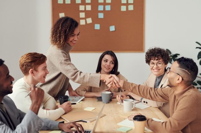 This image shows a group of inviduals at a table of diverse races and ethnicities, sitting at a table scattered with coffee mugs, laptops, notepads, and writing utensils. two individuals shake hands across the table, showing how collaborative discussion creates a positive environment an connects people over new or shared ideas.