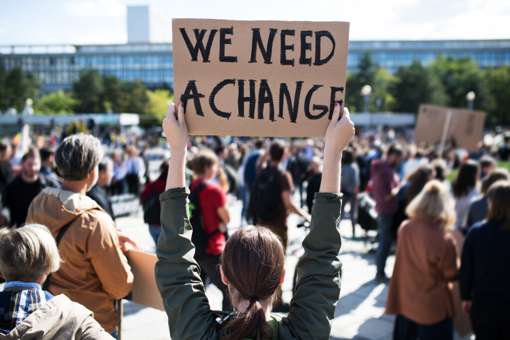 This image focuses on a woman taking part in a protest march, shown from the back, holding up a cardboard poster with the message "WE NEED A CHANGE" in capital letters. This is reminiscent of how citizens are advocating for changes in the existing systems, and need a vehicle for their voice to be heard, supporting Poll the Vote's goals of providing an avenue for anonymous users to voice their concerns via secure polls.