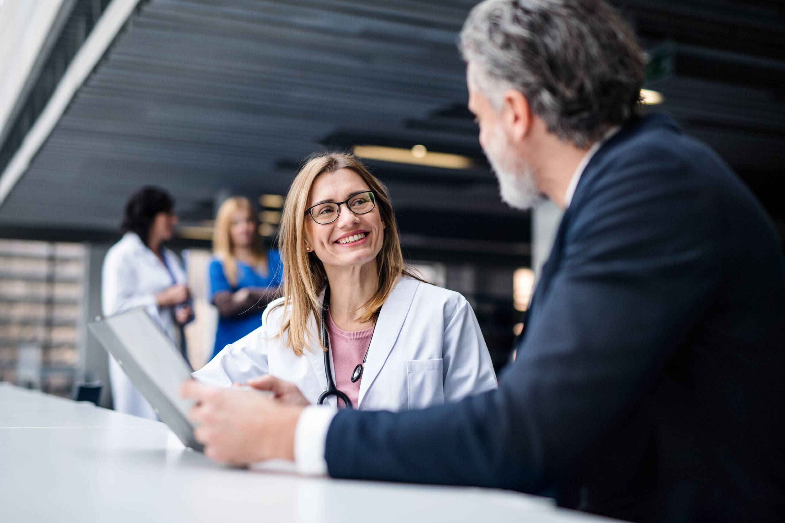 This image portrays a female doctor, wearing glasses and a stethoscope, smiling at a man holding a tablet as they sit in a hospital common room. Her wide smile portrays how healthcare providers create a welcoming and kind environment to assist patients in getting the services and answers that they need.